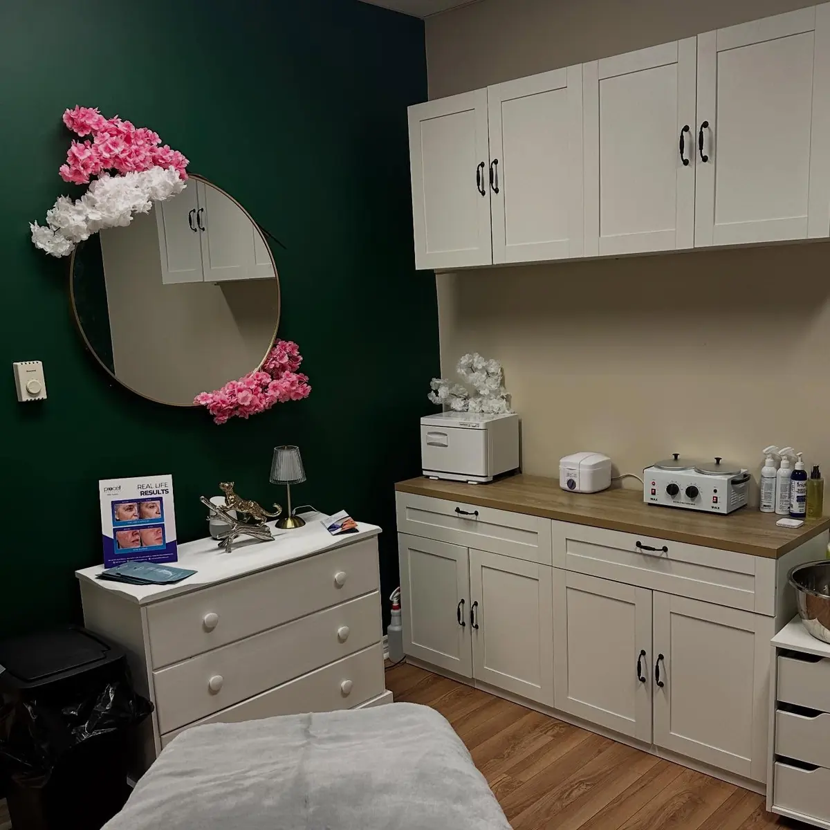 Tori’s treatment room: a deep green wall, a round mirror with pink flowers, white cabinets and a treatment bed
